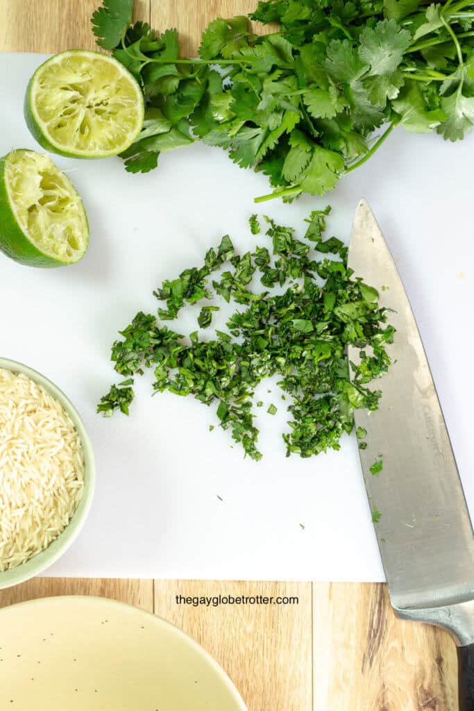 A knife chopping cilantro on a cutting board with limes and rice on it.