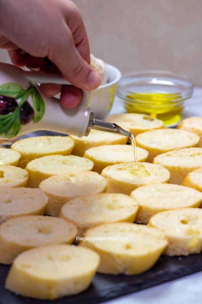 A hand pouring olive oil onto crostini slices.