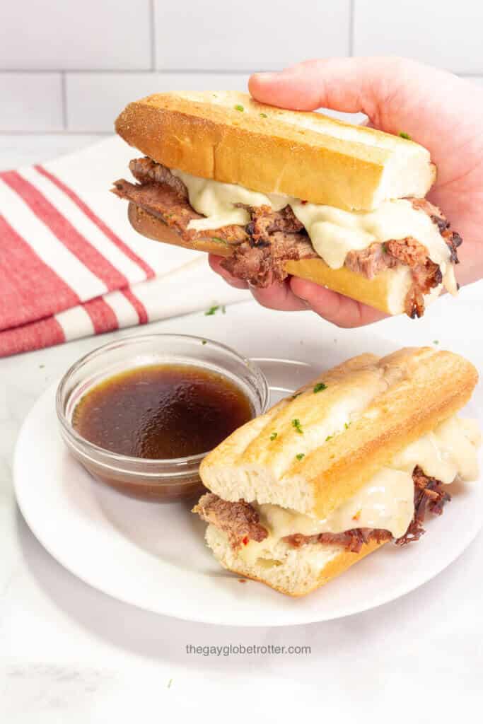 A hand holding a french dip sandwich above a plate with au jus.