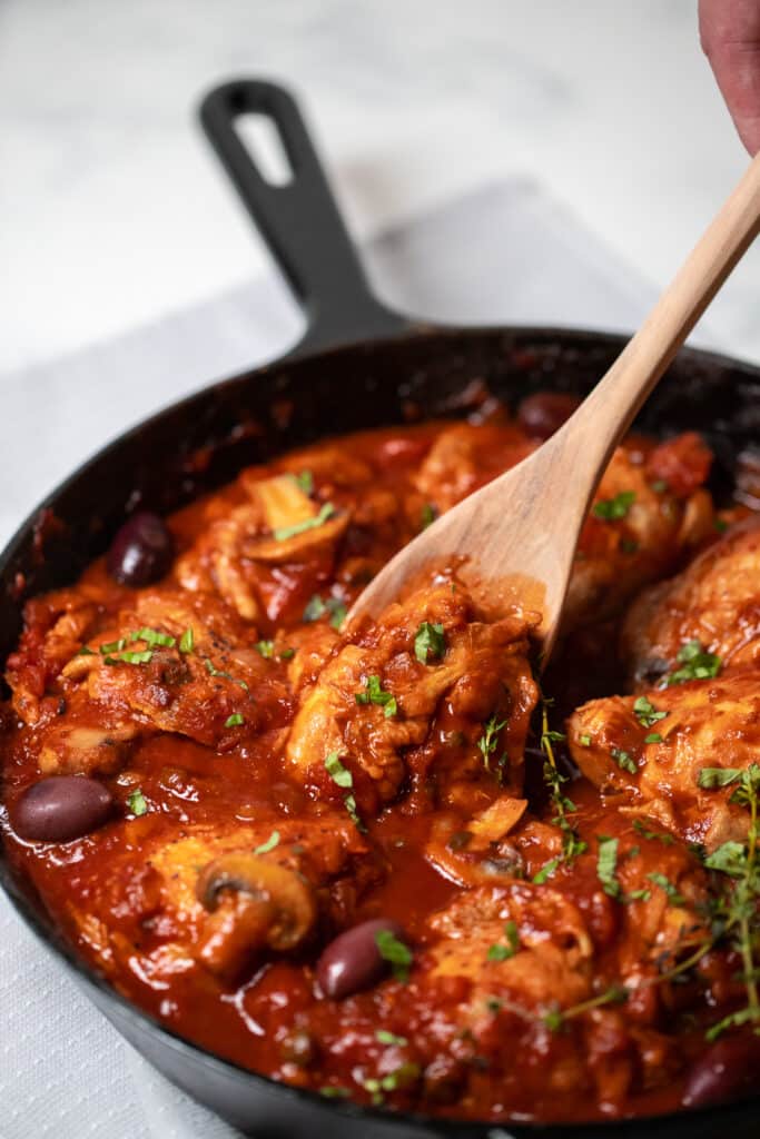 A spoon serving chicken cacciatore from a cast iron pan.