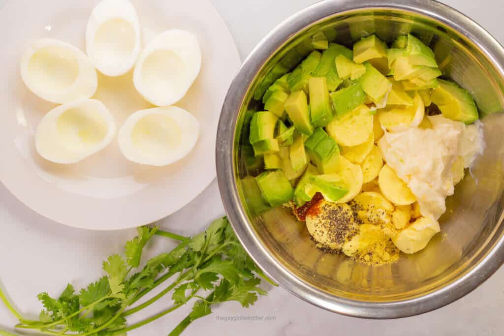 Ingredients in a mixing bowl like egg yolks, avocados, and mayonnaise.