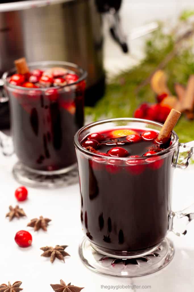 2 cups of slow cooker mulled wine next to a slow cooker, and some pine trees.