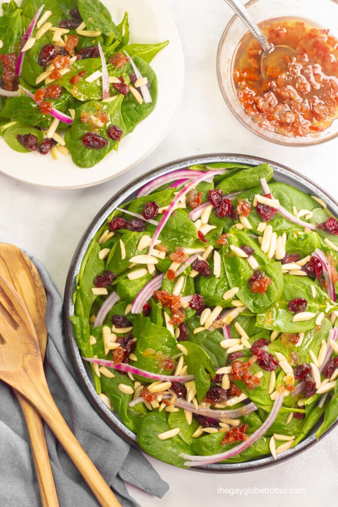 A spinach cranberry salad next to a bowl of warm bacon dressing and serving utensils.