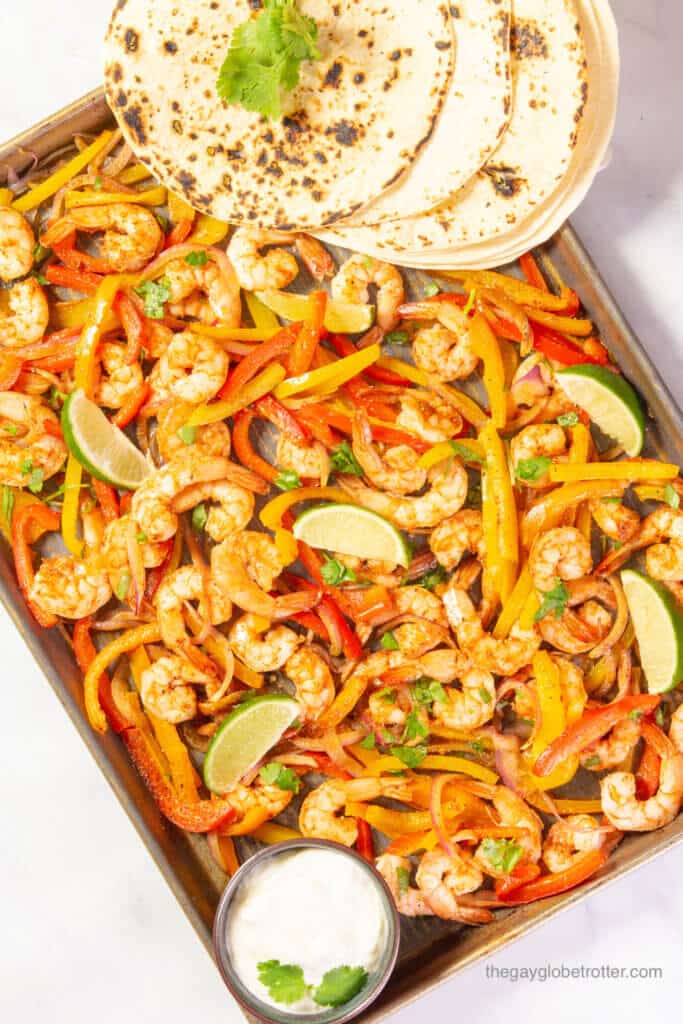 Sheet pan shrimp fajitas with lime wedges, tortillas, and sour cream.