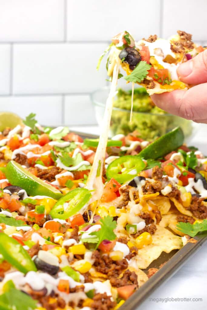A hand grabbing a loaded nacho chip with melted cheese from a sheet pan. 