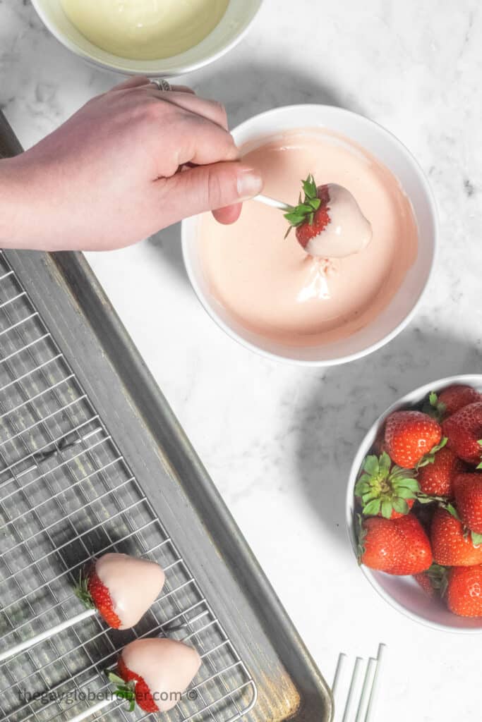 A hand dipping a strawberry into pink chocolate.