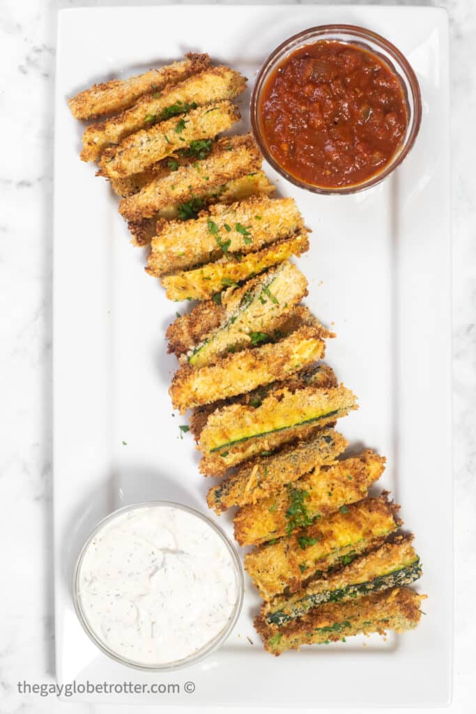 A serving plate of baked zucchini sticks with dill dip and marinara sauce.