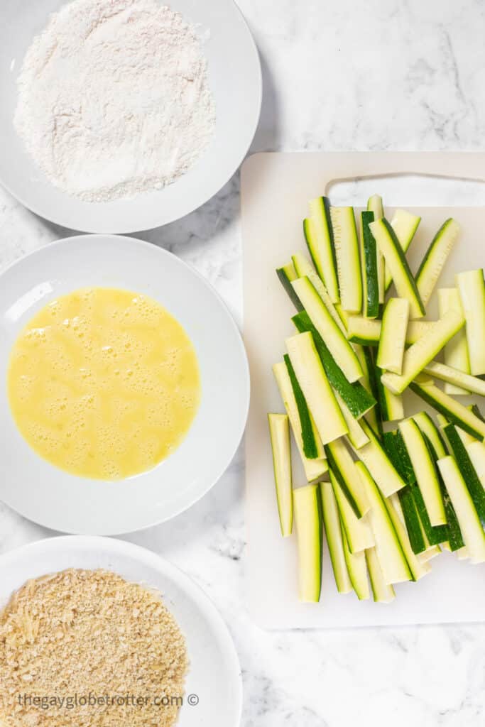 Zucchini sticks next to a bowl of flour, breadcrumbs, and eggwash.