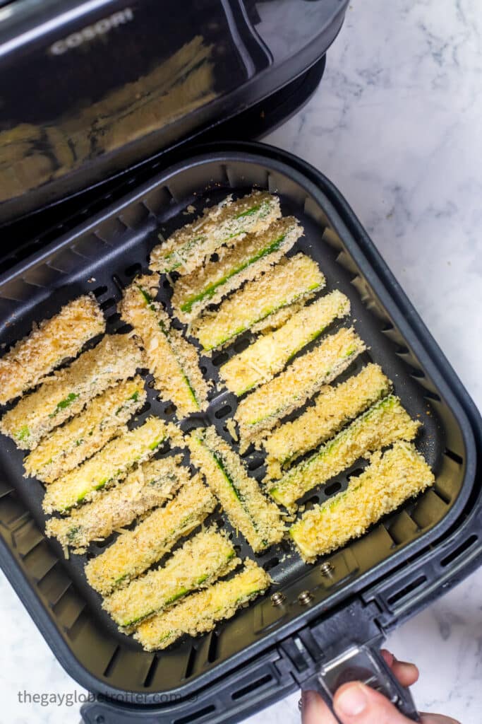 Zucchini fries in an air fryer basket being placed into an air fryer.