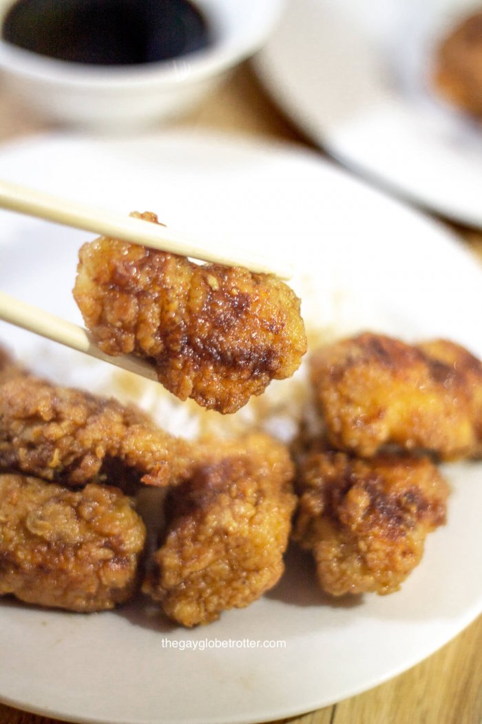 A piece of Korean fried chicken being held by 2 chopsticks with a plate in the background.